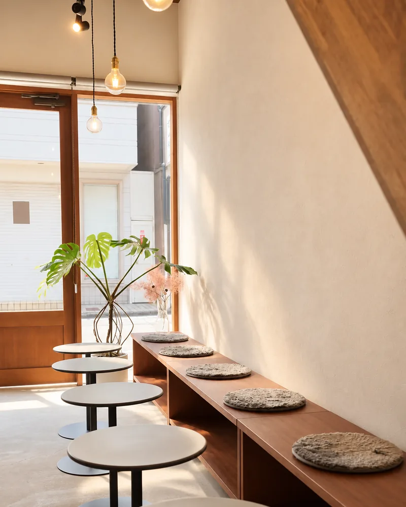 Round tables leading toward the wooden entrance beneath pendant lights, with a cushioned wooden bench running along the wall