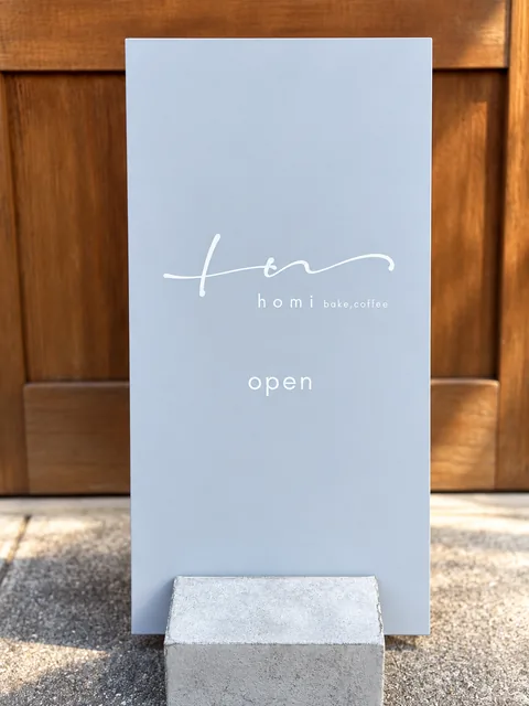 A blue-gray standing sign in front of a wooden door, with a handwritten-style logo, “homi bake,coffee” and “open”