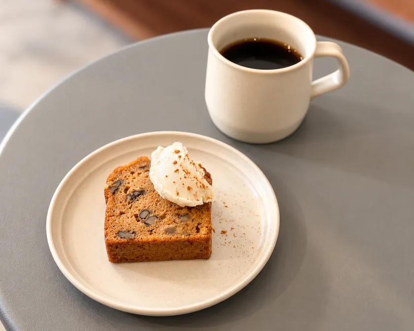 On a blue-gray round table, a plate of carrot cake with cream sits next to a white mug of coffee